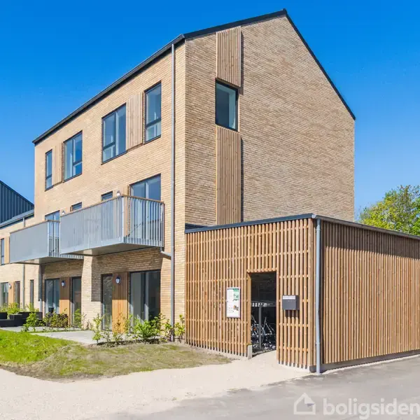 En moderne lejlighedsbygning i tre etager med murstensfacade og balkoner. Ved siden af er der en cykelskur i trædesign. Omkring bygningen er der grønne områder og klar blå himmel.