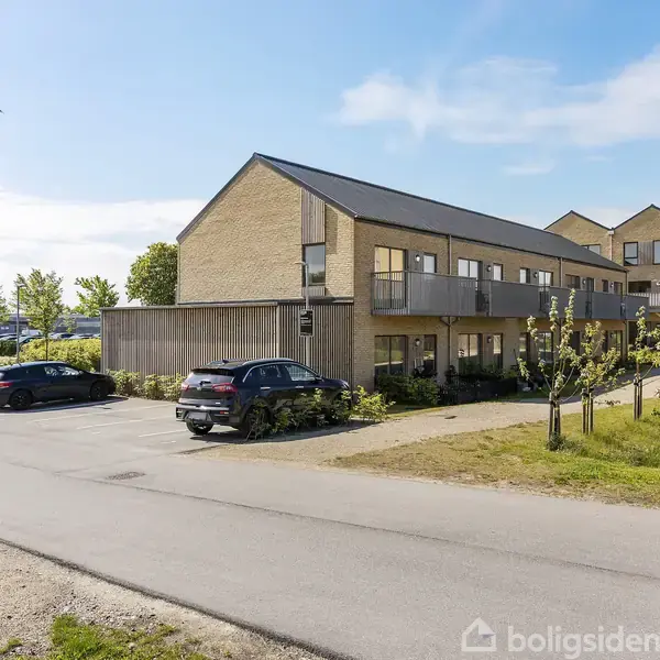 Bygning i to etager med fladt tag. Flere biler parkeret foran på en asfalteret vej. Omkringliggende område med grønne buske og træer under en blå himmel.