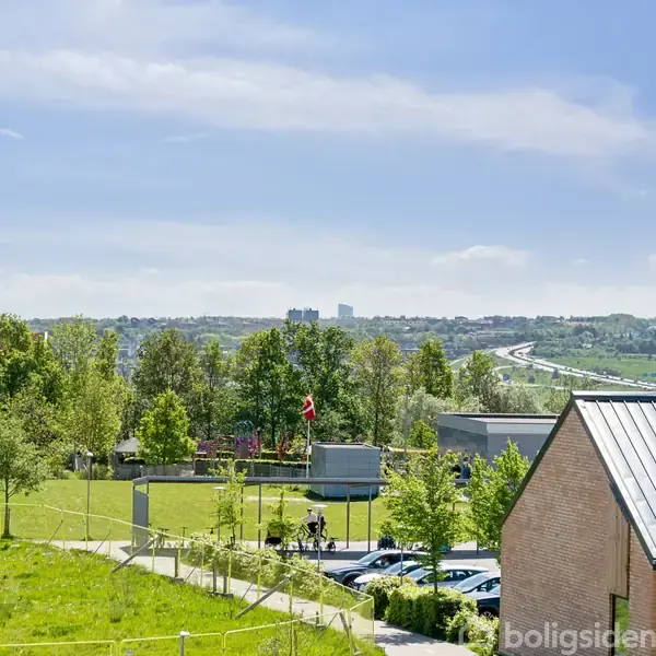 Grøn park med gående mennesker og et lille flag. Parken er omgivet af træer og en bygning med gråt tag. I baggrunden ses en by med moderne bygninger.