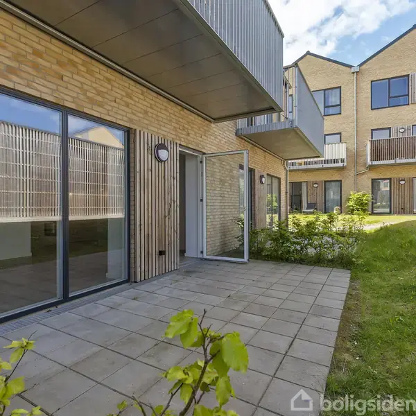 Bygning med store vinduer og altaner, placeret i et grønt område med flisebelagt terrasse i forgrunden. Den blå himmel øger den lyse atmosfære omkring ejendommen.