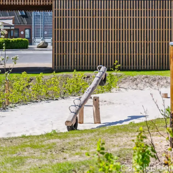 En gynge i træ sidder stationært på sandgrund i en legeplads. Omkranset af grønne planter og hegn med en bygning i baggrunden.