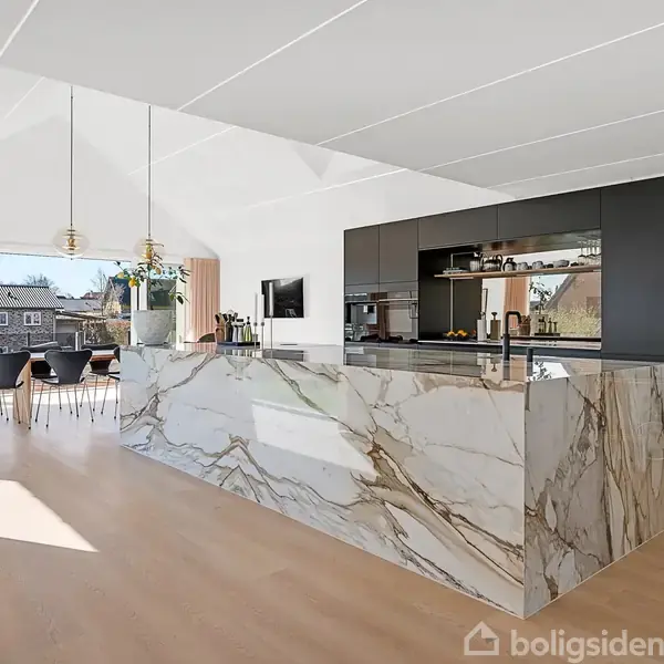 Et moderne køkken med en stor marmorbordplade står i centrum. Spisebord og stole ses i baggrunden ved store vinduer, der giver udsigt til et hus overfor.