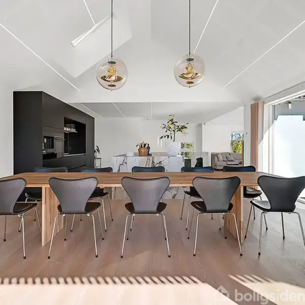 Lang spisebord omgivet af otte stole står i et lyst, moderne køkken-alrum. To pendellamper hænger over bordet, og sorte køkkenelementer ses til venstre. Vinduerne slipper naturligt lys ind.