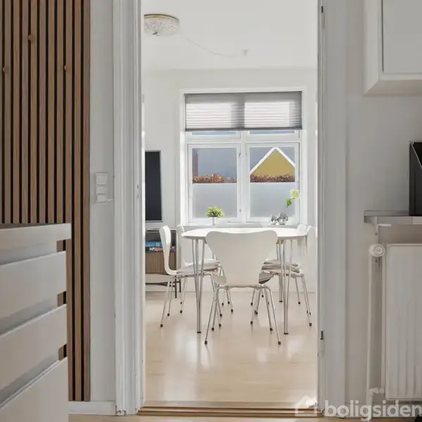 Et lyst spisebord med fire stole står foran et vindue. Udenfor ses en gul husgavl. Rummet er lyst og moderne indrettet med en skærm i baggrunden.