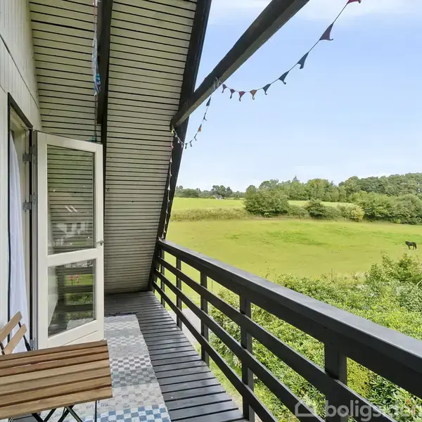 Balkon med træmøbler står stille under en overdækning, pyntet med farverige flag, der afslører en udsigt over grønne marker og træer i baggrunden.