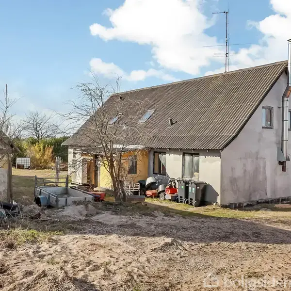 Et hvidt hus med en grå tag hældning står stille ved en grusvej. Omgivet af træer og en træbygning på en solrig dag.