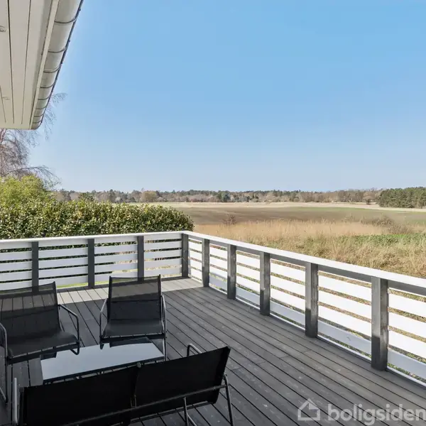 Terrasse med sorte møbler, herunder fire stole omkring et glasbord, mod en baggrund af åbne marker og skov. Sollys fra en klar blå himmel skinner overalt.