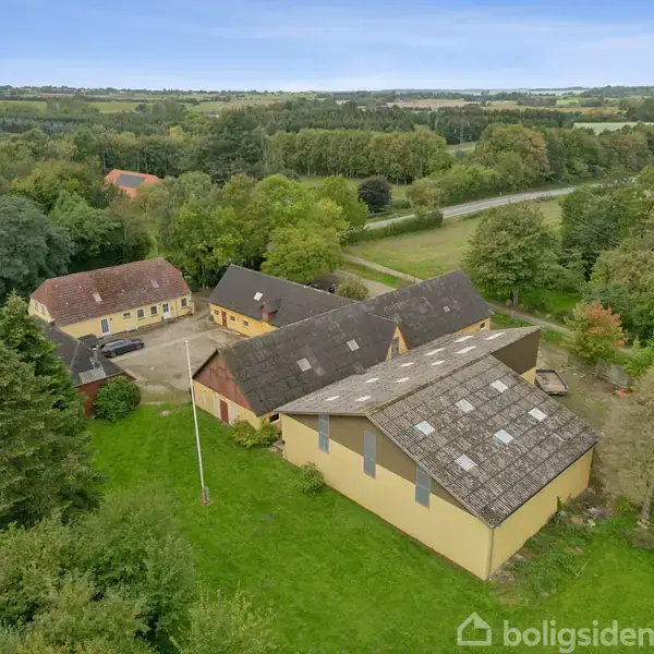 Gule bygninger i gårdkompleks med gårdsplads, omgivet af træer og marker. En stor vej løber i baggrunden, hvilket antyder landlig beliggenhed. Skilt med teksten "boligsiden" nederst til højre.