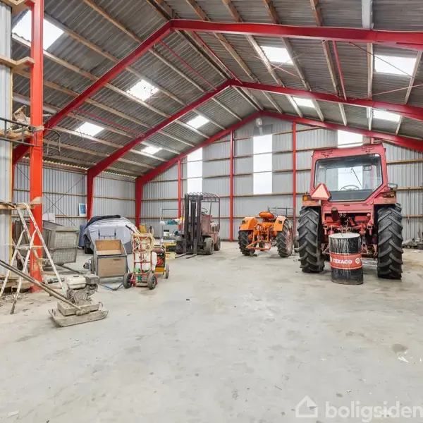 Traktor står stille i en stor lade med metaltag og røde bærende bjælker. Omkring står diverse landbrugsmaskiner, værktøj og materialer spredt på det betonbelagte gulv.
