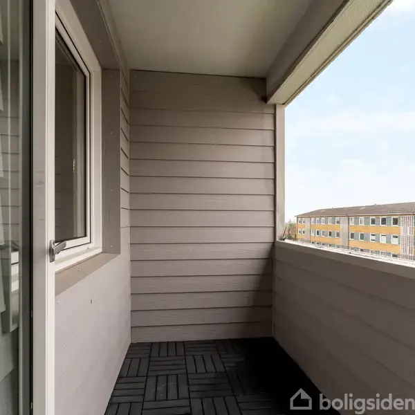 Altan med åbent glasdør, fører til en grå væg med vandret træpaneler. Terrassegulvet er sort. I baggrunden ses boligblokke under en blå himmel.