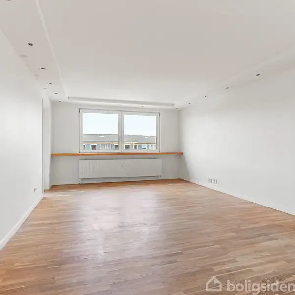 Et tomt værelse har hvidmalede vægge og trægulv. En enkelt radiator står under et stort vindue med udsigt til en bygning. Rummets loft har indbyggede spotlys.
