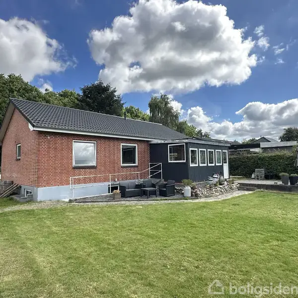Hus med rød murstensfacade og sort tag står i sollys. Foran huset er en terrasse med havemøbler. Græsplæne omgiver huset, himlen er skyet.