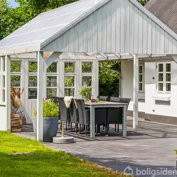 Lyst træpavillon over en spiseplads med sorte stole og bord; placeret på flisebelagt terrasse ved hvidt hus i frodig haveomgivelse.