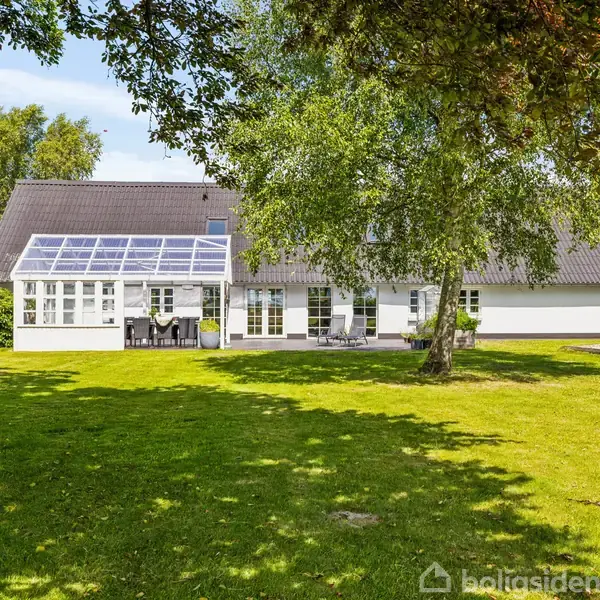 En hvid herregård ligger omgivet af grønne marker og træer. En bil holder parkeret ved siden af bygningen, som er placeret i et landligt landskab under en blå himmel.