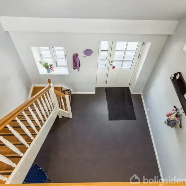 Trappeopgang med trægelænder leder ned til en entré. Døren er omgivet af vinduer. Krog med tøj og en hylde med dekorationer er monteret på væggen.