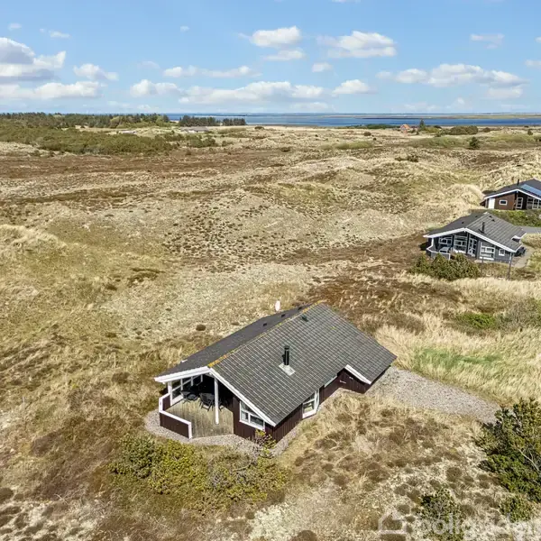 Et træhus med mørkt tag står stille i et sandklitlandskab omkranset af lyng og græs. Andre huse ses i nærheden, og i baggrunden er havet synligt under en blå himmel.