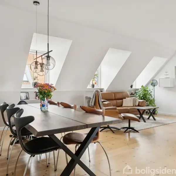 En spisebord med sorte stole står i et lyst rum med skråvinduer. Ved siden af ses en brun sofa og grønne planter. Et moderne, stilrent interiør med trægulv.