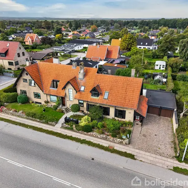 Et stort hus med rødt tegltag står ved en vej. Haven er anlagt med buske og græs. Det er omgivet af andre huse i et grønt, boligområde.