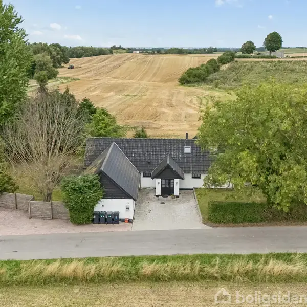 Et hvidt hus med mørkt tag står fredeligt. Det er omgivet af frodige træer og ligger ved en vej med åbne marker i baggrunden, hvilket skaber en landlig atmosfære.