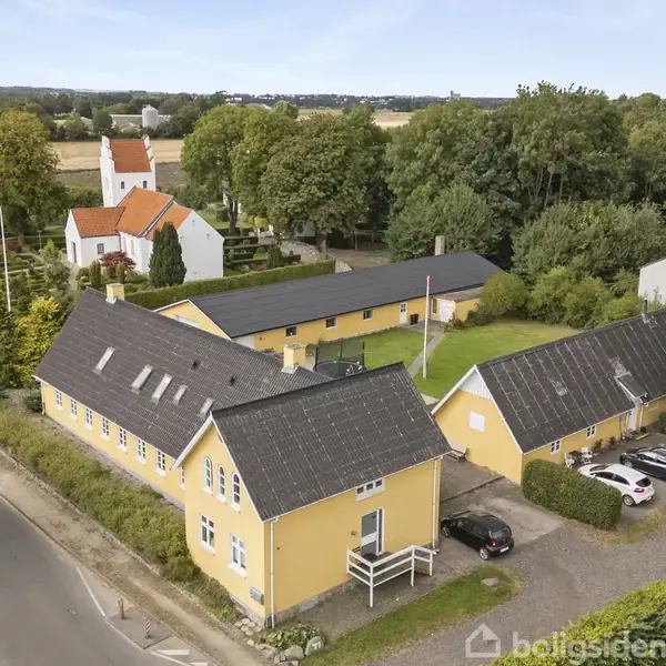 Gule bygninger står stille langs en vej i landlig landsby. Biler er parkeret ved siden af, mens en kirke og træer omgiver området.