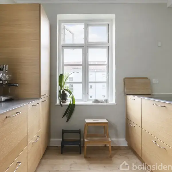 Et lyst køkken med træindretning, espresso-maskine på bordet til venstre, et stort vindue i midten, og en skammel samt plante foran vinduet.