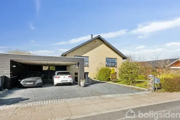 Hus med et skråt tag ligger i et beboelseskvarter. To biler holder parkeret i en dobbelt carport ved siden af huset. Der er grønne buske og en lille have foran.