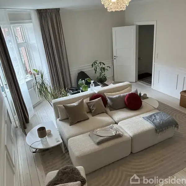 En lys stue med en hvid, komfortabel sofa fyldt med puder. Sofaen står foran et stort vindue med planter, og en rund hvidt bord med krus står ved siden af. En åben dør ses i baggrunden.