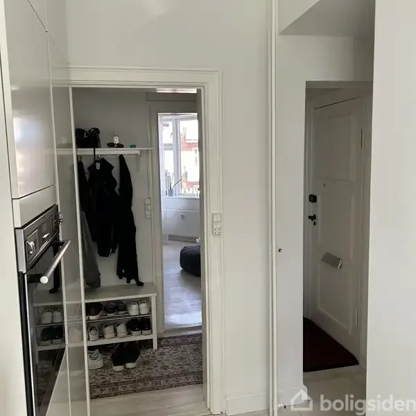 Indgangsområde med knagerække til jakker og hylde til sko. En åben dør fører til et rum med en stor pude ved vinduet. Lyse vægge og træpaneler.
