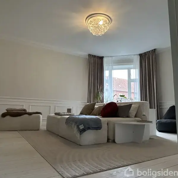 En komfortabel sofa med tæpper og puder står i et lyst stuehjørne. Et vindue med lange gardiner lukker naturligt lys ind i rummet, dekoreret med hvide vægge.