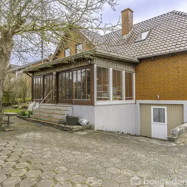 Hus med rød murstensfacade har en glasindfattede veranda. Står i en have med træer og flisebelagt sti. Trappe fører op til verandaen, og der er en kælderdør.