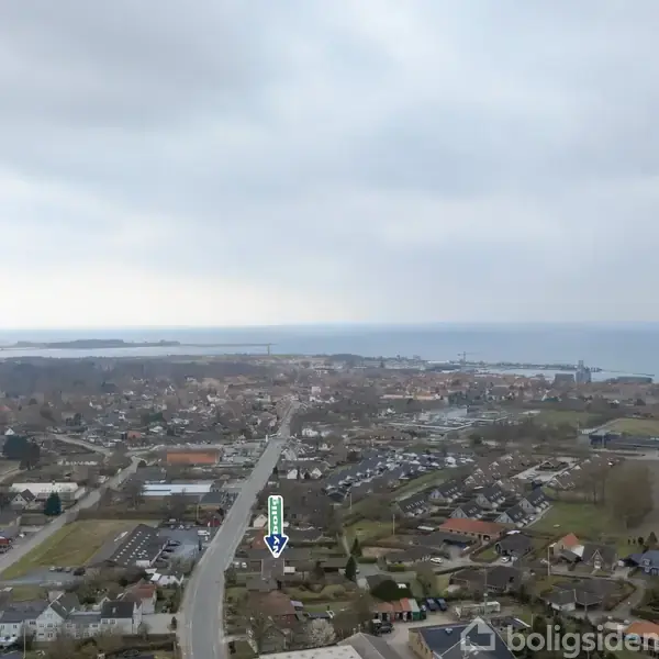 By med adskillige huse ses fra luften. En pil peger ned på et af husene. I horisonten ses havet og en blå himmel med skyer.