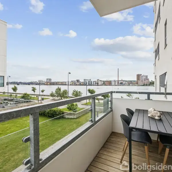 En balkon med et sort bord og seks stole, set mod en udsigt af en flod og moderne bygninger. Grønne områder og flere bygninger omgiver scenen.