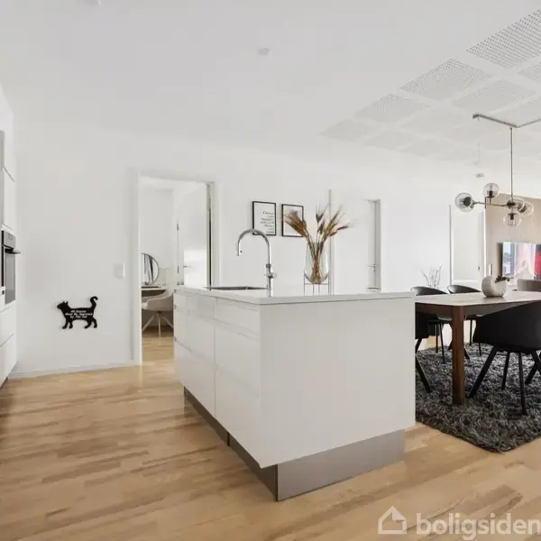 Køkkenø med vask og høje skabe ved siden af spisebord med stole; moderne indretning med lys træparket og vægkunst i et lyst, åbent fællesrum.