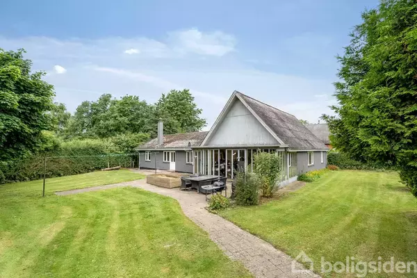 Et gråt hus med gavltag står i en frodig have med velplejet græsplæne og omkranset af træer og buske. En flisebelagt sti fører op til huset.