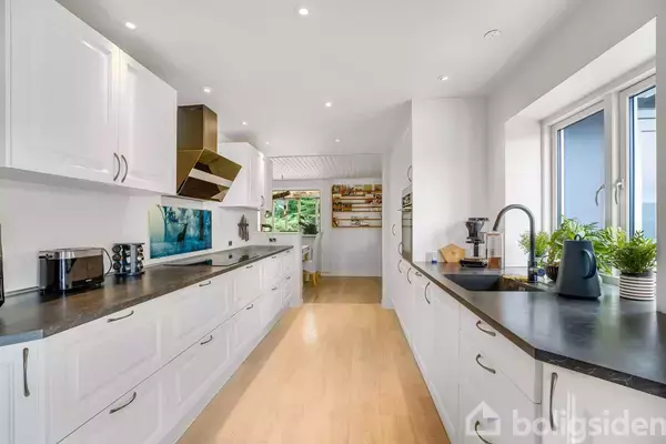 Et moderne køkken med hvide skabe er udstyret med sorte bordplader. Kaffemaskine og planter står ved vinduet, der giver udsigt til en grøn baghave.