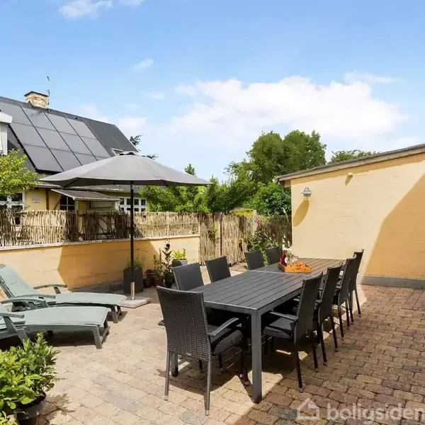 Terrassemøbler står under parasol på en gårdsplads med brolagte sten. Et spisebord med stole dominerer venstre side nær en gul facade. Hække og grønne buske omkranser rummet.