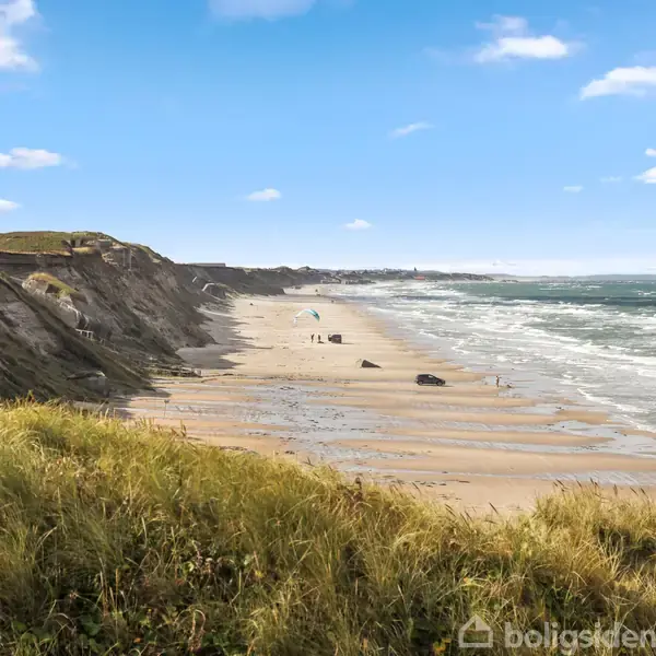 Strand med bølgende hav. Mennesker går langs kystlinjen ved foden af klitter i forgrunden. Blå himmel med spredte skyer danner en klar baggrund.
