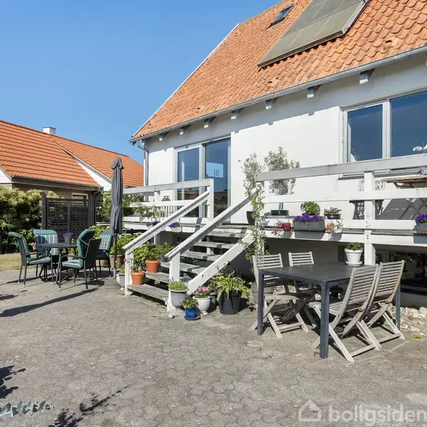 Et hus med rødtag og terrasse, hvor der står et spisebord og stole. Omkring terrassen er der planter og parasoller, og i baggrunden ses et anneks med tilsvarende tag.