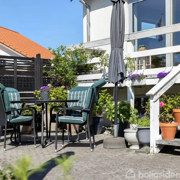 Et sæt havemøbler står på en terrasse med et rundt bord og fire stole. Omkring dem er der blomsterkrukker og en trappe til en hvid bygning.