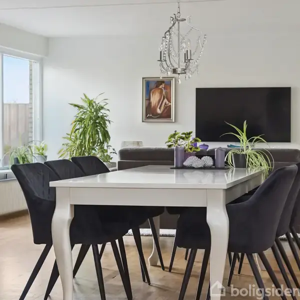 Et hvidt spisebord er omgivet af sorte stole i en stue. Bordet er dekoreret med planter. En lysekrone hænger over bordet. Baggrund: sofa, tv og store vinduer.