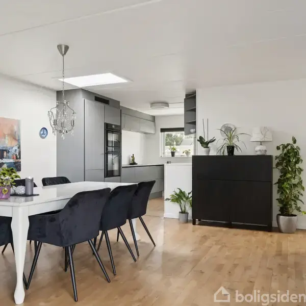 Et moderne spisestuemiljø med en hvid, rektangulær spisebord med sorte stole omkring. Rum med lyse trægulve og hvide vægge, dekoreret med planter og kunst.