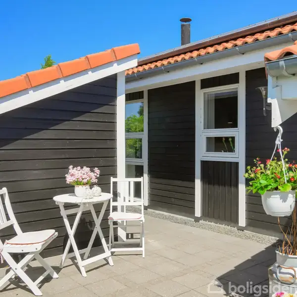Et lille, hvidt cafébord med to stole står på en flisebelagt terrasse foran et sort træhus med rødt tegltag. En blomsterpotte hænger fra taget.