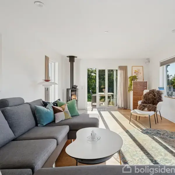 En stue med en grå sofa fyldt med farverige puder står ved et rundt sofabord. En brændeovn tændes i hjørnet. Store vinduer slipper lyset ind.