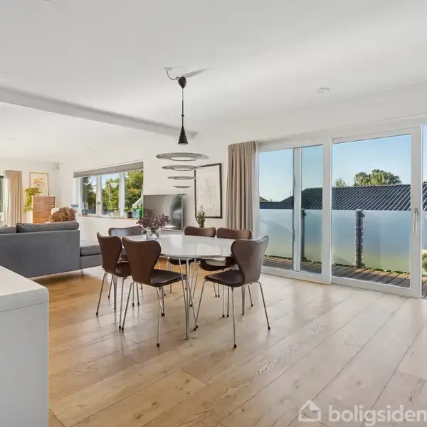 Spisebord med stole står i et lyst rum med trægulv, store vinduer og udsigt til terrasse og grønne omgivelser. En moderne sofa og indretning ses i baggrunden.