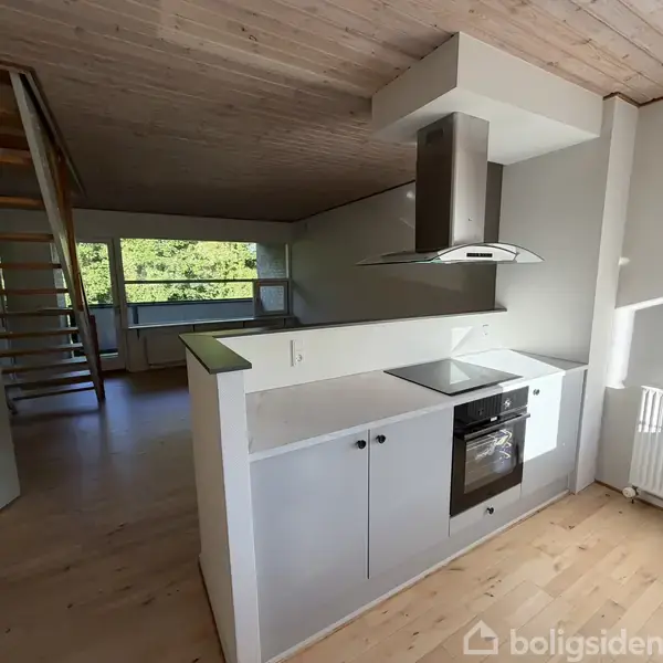 Et lyst køkken med ovn og kogeplade placeret i et åbent rum. En trappe fører op til en anden etage. Vinduer i baggrunden giver udsigt til grønne omgivelser.