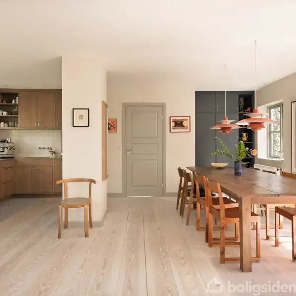 Spisebord omgivet af træstole, placeret i et lyst rum ved siden af et åbent køkken. Dekorativ kunst hænger på væggene, og hængelamper er over bordet.
