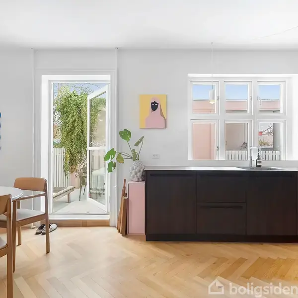 Et lyst køkken-alrum med mørke skabe, hvor en vase med blomster står på et rundt spisebord. Døren fører til en altan med grønne planter. Kunstværker hænger på væggene.