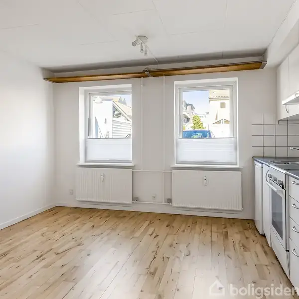Et tomt køkken med trægulv, hvidmalede vægge og to vinduer, der lukker naturligt lys ind. En hvid komfur med ovn er placeret ved siden af et gråmønstret køkkenbord.