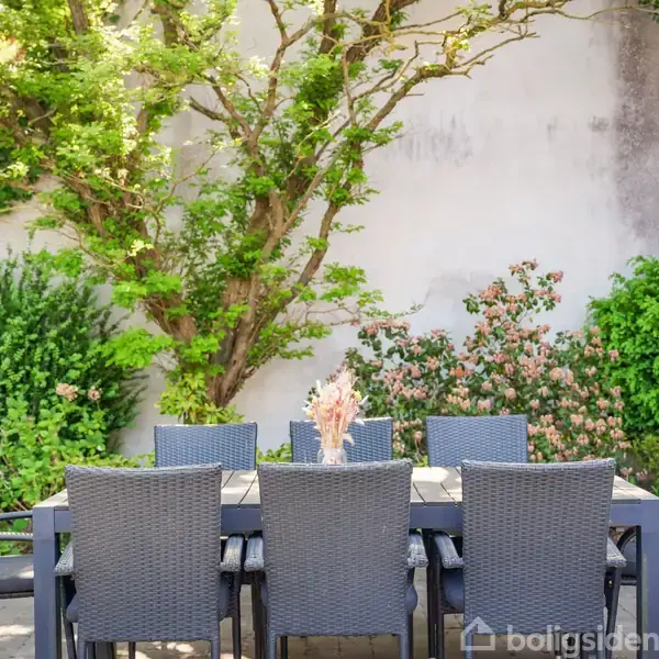Et havebord med otte grå stole står foran en hvid væg. Væggen er delvist dækket af grønne buske og blomstrende planter. En vase med tørrede blomster er på bordet.