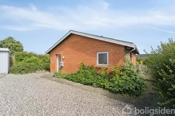 Hus i røde mursten med en hvid dør og vindue til venstre, omgivet af grussti og grønne buske. Skyfri blå himmel i baggrunden.